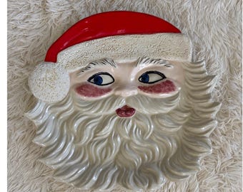 May include: A ceramic Santa Claus face plate with a red hat, white beard, and rosy cheeks. The plate has blue eyes and a white pom-pom on the hat. A festive holiday decoration.