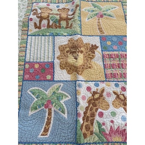 May include: A patchwork quilt with a jungle theme, featuring squares with monkeys, palm trees, a lion, and giraffes. The quilt has a blue, green, yellow, and pink colour scheme with a quilted texture.
