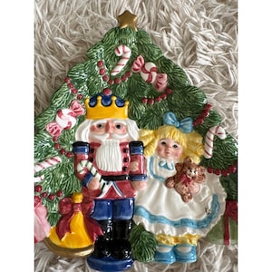 May include: A Christmas tree-shaped ceramic plate featuring a nutcracker soldier and a girl holding a teddy bear. The plate is decorated with candy canes, a gold star, and a bell. The figures are in bright colors.