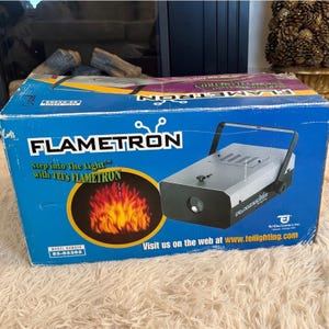 Peut inclure: Une boîte bleue avec le mot "FLAMETRON" en lettres blanches. La boîte présente un graphique de flammes et un appareil argenté avec une poignée. Le texte "Step into The Light" est également visible.