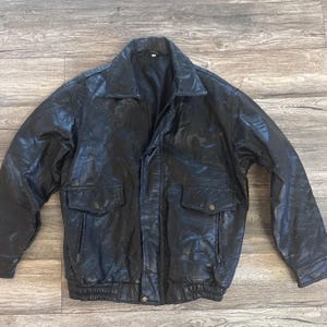 May include: Black leather jacket with a collared neckline, two front pockets with button closures, and two side pockets with zippers. The jacket has a ribbed hem and cuffs.