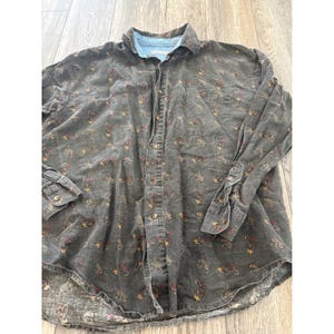 Puede incluir: Camisa de manga larga gris oscuro con un estampado floral en tonos rojos, amarillos y marrones. La camisa tiene cuello y puños azul claro. Botones dorados.