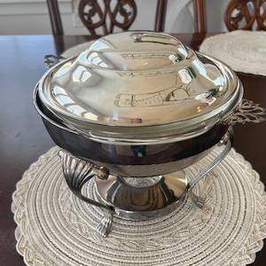 Könnte beinhalten: Ein silberfarbenes Chafing Dish mit gewölbtem Deckel und dekorativen Griffen, das auf einem dekorativen Ständer steht. Das Gericht steht auf einem runden, gewebten Platzset. Die reflektierende Oberfläche spiegelt die Umgebung wider.