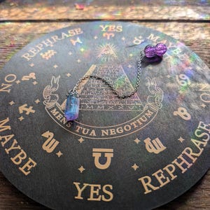 Könnte beinhalten: Eine schwarze und goldene Planchette mit den Wörtern "YES", "MAYBE" und "REPHRASE" aufgedruckt. Ein Kristallpendel hängt über der Planchette. Das Pendel hat einen blauen und lila Kristall und eine lila Perle.
