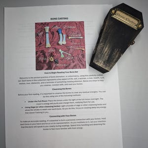 Op de afbeelding: Een zwarte, doodskistvormige doos met een gouden label met de tekst "Bone Casting" ligt naast een bedrukte pagina. De pagina bevat een diagram van botvormen en de woorden "Bone Casting" en instructies.