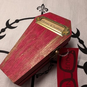 Op de afbeelding: Een kleine, roze houten kist met gouden accenten en een messing naamplaatje met de tekst "Bone Casting". Een rood label met de woorden "FOUR NERD" en een brilgrafiek hangt aan de kist.