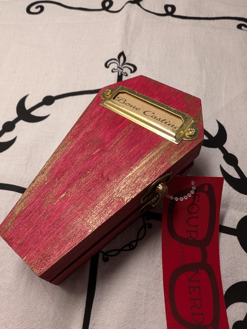 Op de afbeelding: Een kleine, roze houten kist met gouden accenten en een messing naamplaatje met de tekst "Bone Casting". Een rood label met de woorden "FOUR NERD" en een brilgrafiek hangt aan de kist.