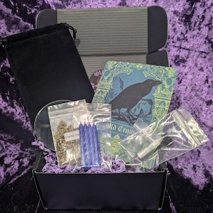 May include: A black box filled with various items, including a black velvet bag, a small mirror, and several small plastic bags containing herbs and blue candles. A card with a raven illustration and the text "Old Crow" is also visible.