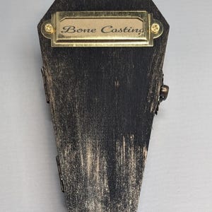 Op de afbeelding: Een doodskistvormige houten kist met een verweerde zwarte en gouden afwerking. De kist heeft een messing naamplaatje met de tekst "Bone Casting". Scharnieren en een sluiting zijn zichtbaar aan de zijkanten. Gothic of Halloween thema.