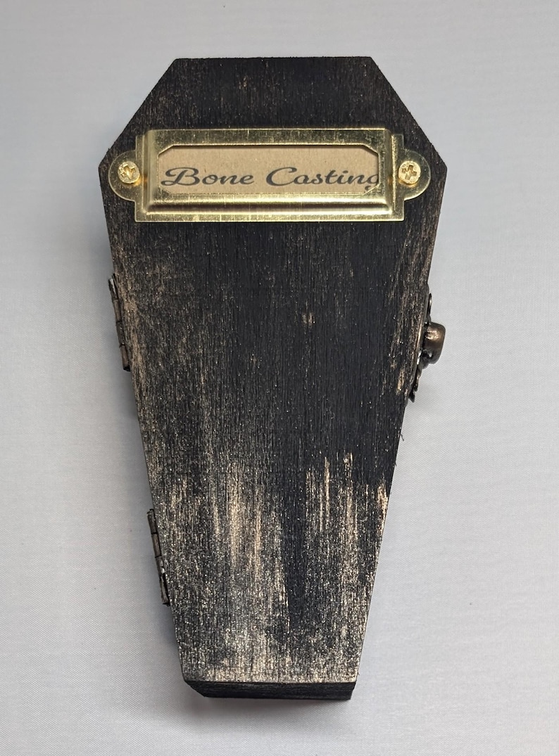 Op de afbeelding: Een doodskistvormige houten kist met een verweerde zwarte en gouden afwerking. De kist heeft een messing naamplaatje met de tekst "Bone Casting". Scharnieren en een sluiting zijn zichtbaar aan de zijkanten. Gothic of Halloween thema.