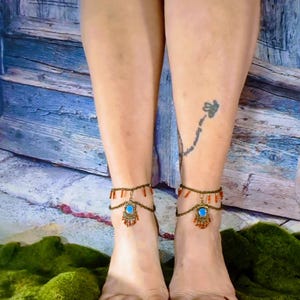 May include: Anklets with a bronze chain and decorative elements. Each anklet features a central turquoise-colored stone, surrounded by smaller orange beads. The anklets are displayed on a person's feet, with a tattoo visible on the leg.