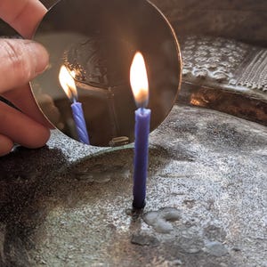 Könnte beinhalten: Eine brennende blaue Kerze mit einer hellgelben Flamme spiegelt sich in einer kreisförmigen, dunklen Metalloberfläche. Die Kerze steht auf einer strukturierten, grauen Oberfläche, mit Wachs um ihren Fuß. Eine Hand hält die reflektierende Oberfläche.