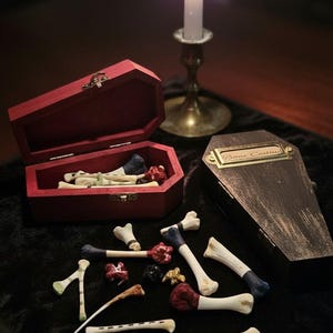 Op de afbeelding: Een donkere compositie met een brandende kaars, miniatuur doodskisten en botvormige objecten. Een rode doodskist is open en toont botten en rode details. De andere doodskist heeft een messing naamplaat met de tekst "Bone Casting".