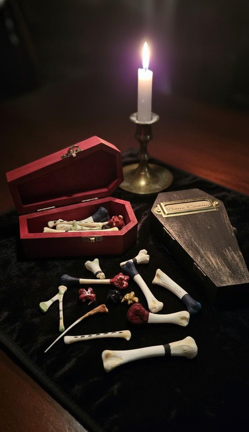 Op de afbeelding: Een donkere compositie met een brandende kaars, miniatuur doodskisten en botvormige objecten. Een rode doodskist is open en toont botten en rode details. De andere doodskist heeft een messing naamplaat met de tekst "Bone Casting".