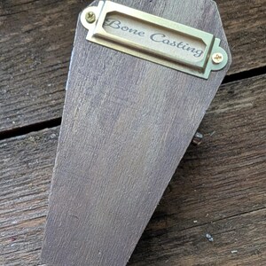 Op de afbeelding: Een houten kistvormige doos met een messing labelhouder met de tekst "Bone Casting". De doos is gemaakt van gebeitst hout en heeft een kleine messing knop. De achtergrond is een houten oppervlak.