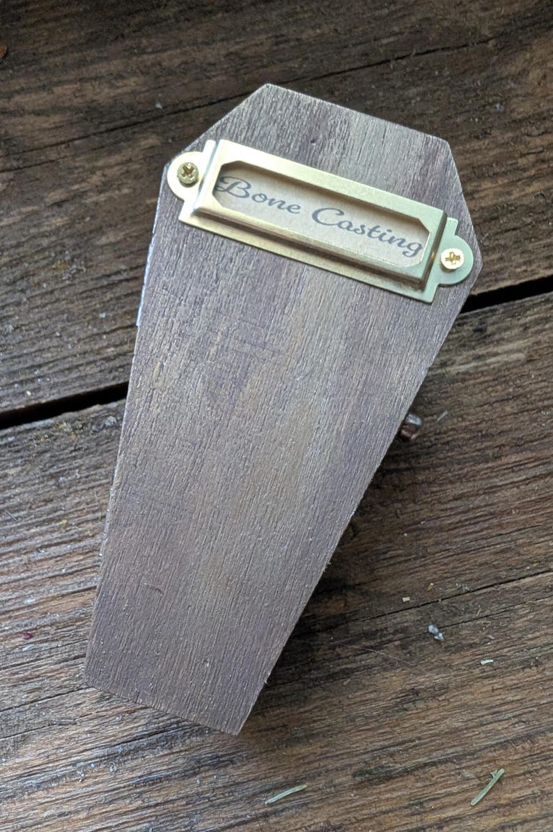 Op de afbeelding: Een houten kistvormige doos met een messing labelhouder met de tekst "Bone Casting". De doos is gemaakt van gebeitst hout en heeft een kleine messing knop. De achtergrond is een houten oppervlak.
