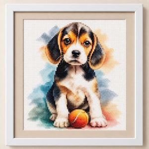 May include: A framed cross-stitch embroidery featuring a beagle puppy with black, brown, and white fur sitting down. The puppy has large dark eyes and is positioned behind a bright orange tennis ball. The background has soft watercolour-style splashes.