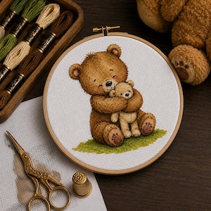 May include: A circular wooden embroidery hoop containing white Aida fabric with a cross-stitch design of a large brown teddy bear hugging a smaller light tan teddy bear, sitting on a patch of green grass, accompanied by embroidery floss and scissors.