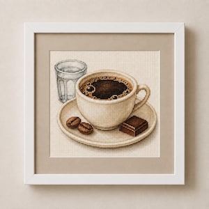 Puede incluir: Un bordado de punto de cruz en un marco blanco que representa una taza de cerámica color crema llena de café negro sobre un platillo a juego. Dos granos de café marrones y un trozo de chocolate negro descansan sobre el platillo junto a un vaso de agua.