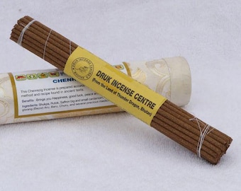 May include: A bundle of brown incense sticks tied with white string, with a yellow label that reads "DRUK INCENSE CENTRE" and text below. A cylindrical container with text is in the background.