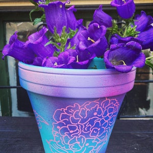 May include: A purple flower arrangement in a blue and pink painted terracotta pot with a pink floral design.