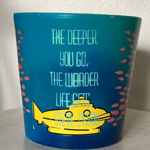 May include: A cylindrical, blue planter with the text "THE DEEPER YOU GO, THE WEIRDER LIFE GETS" in white. A yellow submarine graphic is featured below the text. Small red fish are painted on the sides. The planter is approximately 5 inches tall.