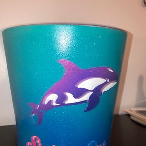 May include: A teal blue and purple ombre ceramic cup with a white and purple orca whale graphic and the text "Sink the Rich" in rainbow colors.