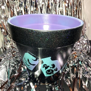May include: Black planter pot with a purple rim and a glittery finish. The pot has a design of two jars with ghosts and bats inside.