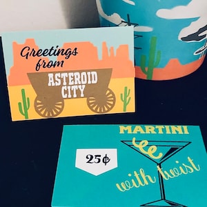 May include: Two paper cards with a western theme. The first card is light blue with a brown wagon and the text "Greetings from Asteroid City". The second card is turquoise with a martini glass and the text "MARTINI with twist" and "25¢".