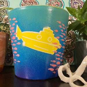 May include: A cylindrical ceramic planter with a gradient blue background. A yellow submarine graphic is featured on the front, surrounded by pink fish. The planter is designed for indoor or outdoor use and is suitable for various plants.