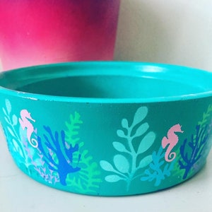 May include: A teal-colored ceramic planter with a seahorse and coral design. The planter is decorated with a light blue coral, pink seahorses, and green seaweed.