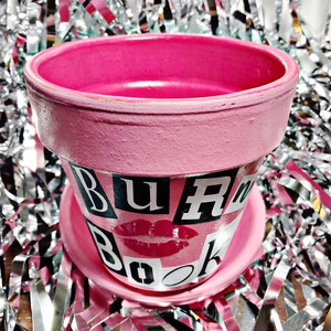 May include: Pink ceramic planter with a white saucer. The planter has the words "BURR Book" in black letters with a red lipstick kiss in the center.