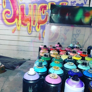 May include: A collection of spray paint cans in various colours, including blue, green, pink, and purple. The cans are arranged in rows on a white surface. The word "JUICY" is painted in pink, yellow, and orange on a wall in the background.