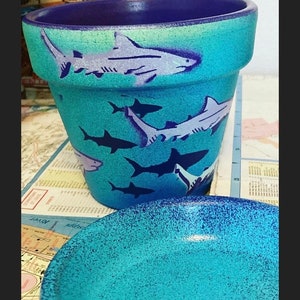 May include: A turquoise ceramic flower pot with a matching saucer. The pot is decorated with white and navy blue shark illustrations. The rim is a darker shade of blue.