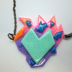 May include: A colorful, geometric heart pendant necklace. The pendant features a layered design with shades of pink, orange, purple, and teal, accented with glitter. The necklace has a dark chain and clasp.