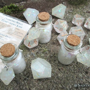 May include: A set of iridescent white dice and two small glass jars filled with iridescent glitter. The jars have cork stoppers and are tied with twine. The dice are numbered 1-20 and have a copper metallic finish. A printed paper with the text "DIAMOND SPELL COMPONENT LIST" is visible in the background.