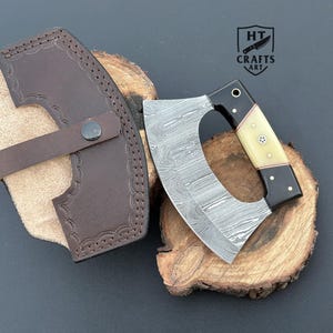 May include: A Damascus steel cleaver knife with a curved blade and a handle with black and cream accents. The knife rests on a wooden surface next to a brown leather sheath. The sheath has a strap and is decorated with a stitched pattern. The logo "HT Crafts Art" is visible.