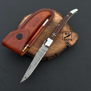 May include: A Damascus steel folding knife with a dark wood handle and silver accents. The knife is open, revealing the patterned blade. A brown leather sheath and a pen are next to the knife on a wooden surface. The logo "HT Crafts Art" is visible.