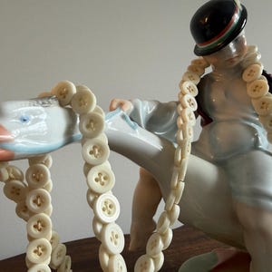 May include: A porcelain figurine of a figure wearing a black bowler hat with a green and red band, riding a large white goose with an orange beak. A long string of cream-colored, four-hole buttons is draped over the figure and the goose.