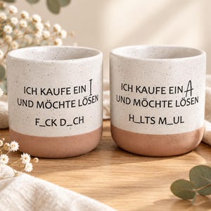 Könnte beinhalten: Zwei Keramikbecher mit weiß gesprenkeltem Oberteil und terrakottafarbenem Boden. Beide Becher tragen den schwarzen Text 'ICH KAUFE EIN' und 'UND MÖCHTE LÖSEN'. Der linke Becher zeigt 'F_CK D_CH', der rechte 'H_LTS M_UL' mit den Buchstaben I und A.