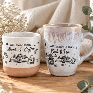 Könnte beinhalten: Zwei Keramikbecher mit gesprenkelter Oberfläche. Der linke Becher zeigt den Text All I need is my Book & Coffee mit einer Buch- und Kaffeetassenillustration. Der rechte Becher hat einen tropfenden Glasurrand und den Text All I need is my Book & Tea.