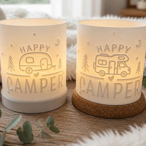 May include: Two cylindrical white lamps with warm internal lighting. Each lamp features embossed text reading HAPPY CAMPER, along with line art illustrations of a camper trailer or motorhome, pine trees, stars, and a crescent moon.
