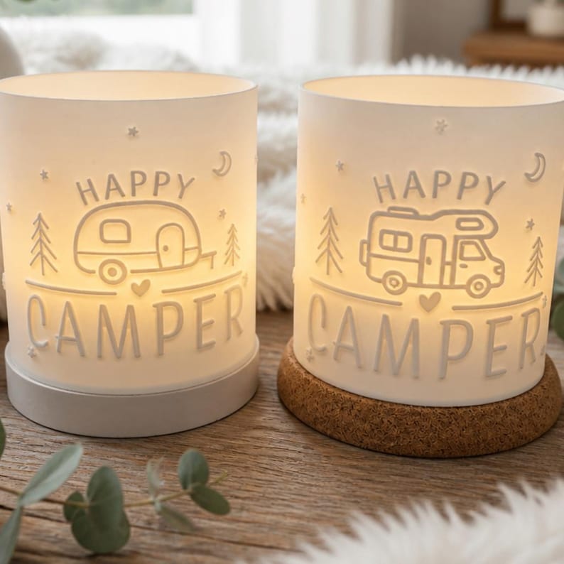 May include: Two cylindrical white lamps with warm internal lighting. Each lamp features embossed text reading HAPPY CAMPER, along with line art illustrations of a camper trailer or motorhome, pine trees, stars, and a crescent moon.