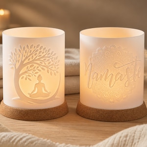 Könnte beinhalten: Zwei weiße Keramik-Kerzenhalter mit Korksockel. Einer zeigt einen Baum und eine meditierende Person, der andere das Wort "Namaste" mit einem Mandala-Design. Die Kerzenhalter sind von innen beleuchtet.