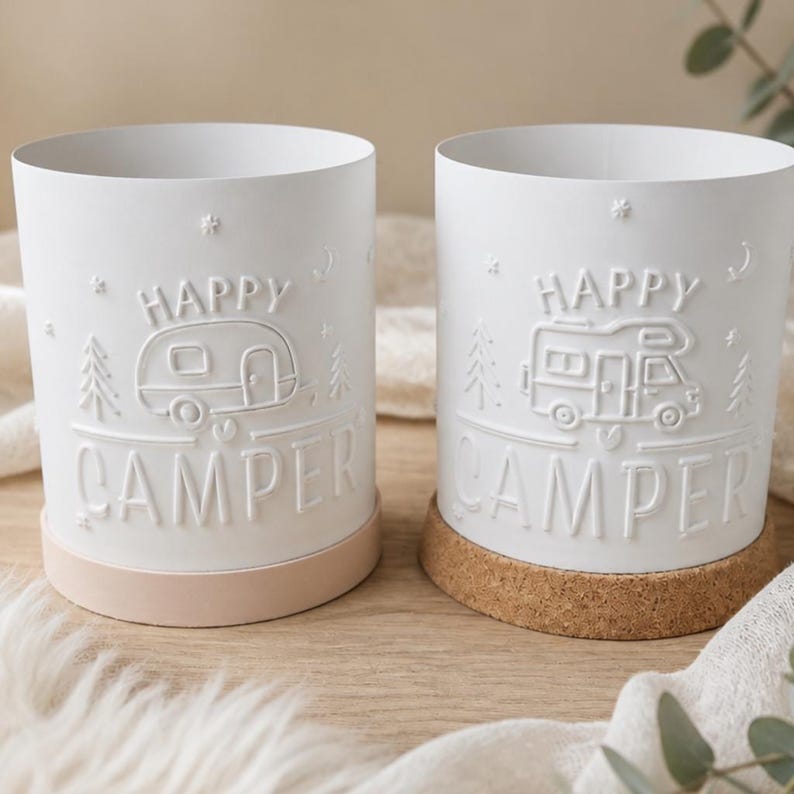 May include: Two white cylindrical containers featuring embossed designs. The left container shows a travel trailer with the text HAPPY CAMPER and a light-coloured base. The right container shows a motorhome with the text HAPPY CAMPER and a cork base.