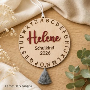 May include: A circular light-colored wooden ornament featuring the alphabet engraved around the perimeter. The name Helene is displayed in raised dark red script lettering, with the text Schulkind 2026 below it. A grey tassel hangs from the bottom.