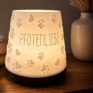 Könnte beinhalten: Ein weißer, konischer, beleuchteter Lampenschirm mit ausgestanzten Pfotenabdrücken und Herzen sowie dem mittig platzierten deutschen Schriftzug "PFOTENLIEBE". Die Lampe hat einen soliden schwarzen runden Sockel und strahlt ein warmes Licht aus.