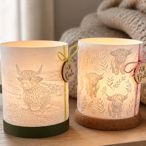 Könnte beinhalten: Zwei zylindrische Kerzenhalter mit Highland-Kuh-Designs. Einer zeigt eine einzelne Kuh, der andere mehrere Kühe und Blättermuster. Jeder hat ein Holzschild mit der Aufschrift "Für Dich". Die Kerzenhalter sind weiß mit grünem und braunem Sockel.