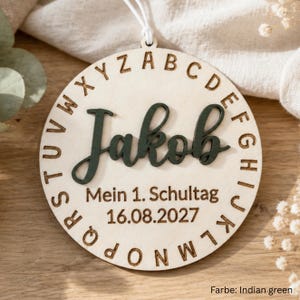 May include: A circular wooden ornament featuring the name Jakob in dark green script, surrounded by an engraved alphabet border. Below the name, the text reads Mein 1. Schultag and the date 16.08.2027. A white string is attached to the top.