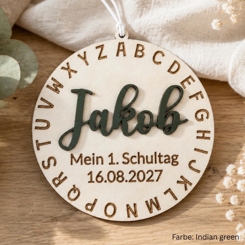 May include: A circular wooden ornament featuring the name Jakob in dark green script, surrounded by an engraved alphabet border. Below the name, the text reads Mein 1. Schultag and the date 16.08.2027. A white string is attached to the top.